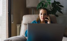 Доросла жінка з ноутбуком, що розмовляє смартфоном під час роботи вдома — стокове фото