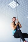 From below side view of young athletic male in sportswear performing rope climb exercise during fitness training in modern gym — Stock Photo