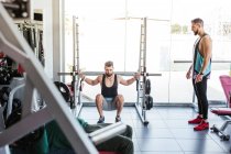 Potente deportista determinado haciendo ejercicio con barra en el bastidor de sentadilla durante el entrenamiento de levantamiento de pesas con entrenador personal en el gimnasio moderno - foto de stock