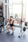 Potente deportista determinado haciendo ejercicio con barra en el bastidor de sentadilla durante el entrenamiento de levantamiento de pesas con entrenador personal en el gimnasio moderno - foto de stock