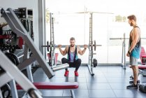 Esportista determinado poderoso fazendo exercício com barra no rack de agachamento durante o treinamento de levantamento de peso com treinador pessoal no ginásio moderno — Fotografia de Stock