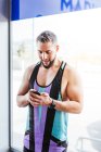 Homem musculoso excitado em sportswear com foco na tela e interagindo com o smartphone com prazer enquanto está sozinho contra a parede de vidro no corredor do ginásio contemporâneo — Fotografia de Stock