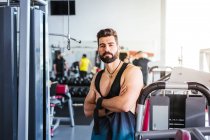 Muskulöser bärtiger Mann in Sportbekleidung, der mit verschränkten Armen in die Kamera blickt und sich in einem leichten modernen Fitnessstudio an Sportgeräte lehnt — Stockfoto