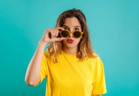 Junge Frau im gelben Outfit blickt in die Kamera und passt stylische Sonnenbrille vor buntem türkisfarbenem Hintergrund an — Stockfoto