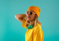 Seitenansicht einer fröhlichen jungen hübschen Frau in gelbem Outfit und orangefarbener Mütze, die mit Sonnenbrille vor buntem türkisfarbenem Hintergrund wegschaut — Stockfoto