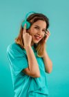 Side view of happy young woman in bright t shirt smiling looking at camera and listening to music in earphones against turquoise background — Stock Photo