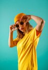 Joven hembra en traje amarillo mirando a la cámara y ajustando elegantes gafas de sol sobre fondo turquesa - foto de stock