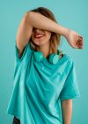 Щаслива молода жінка в яскравій сорочці посміхається рукою над обличчям, прикриваючи очі навушниками, що відпочивають на її шиї на бірюзовому фоні — стокове фото
