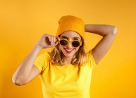 Счастливая молодая женщина в яркой одежде и стильных солнечных очках улыбается и смотрит в камеру на оранжевом фоне — стоковое фото