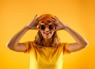 Jovem feliz em roupas brilhantes e óculos de sol elegantes sorrindo e olhando para a câmera contra o fundo laranja — Fotografia de Stock