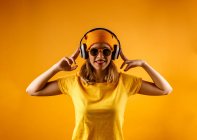 Glückliche junge Frau in heller Kleidung und stylischer Sonnenbrille lächelt in die Kamera, während sie Musik vor orangefarbenem Hintergrund hört — Stockfoto