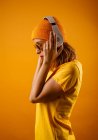 Glückliche junge Frau in heller Kleidung, die stylische Sonnenbrille aufsetzt und weglächelt, während sie Musik vor orangefarbenem Hintergrund hört — Stockfoto