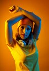Уверенная в себе стильная молодая женщина в оранжевой шапочке с наушниками вокруг шеи, держащая руку над головой и глядя на камеру на оранжевом фоне — стоковое фото