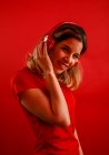 Vue latérale d'une jeune femme blonde heureuse avec des écouteurs vifs et un t-shirt souriant et regardant la caméra tout en écoutant de la musique sur fond rouge — Photo de stock