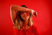 Happy young woman smiling and looking at camera while standing against red background with arm over the head — Stock Photo