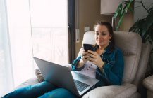 Reife Frau mit Laptop surft Smartphone und schaut weg, während sie im Sessel sitzt und zu Hause an einem Remote-Projekt arbeitet — Stockfoto