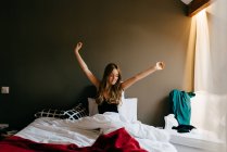 Щаслива мокра молода жінка в сплячому одязі, що піднімає руки із закритими очима, розтягуючи тіло, сидячи на зручному ліжку після пробудження вдома — стокове фото