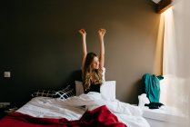 Счастливая спокойная молодая женщина в сонной одежде поднимает руки с закрытыми глазами растягивая тело, сидя на удобной кровати после пробуждения утром дома — стоковое фото