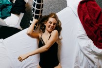Draufsicht der aufgeregten schönen jungen Frau in Unterwäsche, die lachend in die Kamera blickt und Kissen wirft, während sie morgens zu Hause auf einem bequemen Bett liegt — Stockfoto