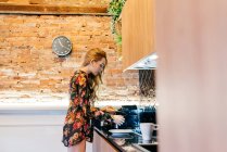 Vista lateral de la joven mujer feliz en blusa floral poniendo pan en tostadora mientras cocina el desayuno en la acogedora cocina por la mañana - foto de stock