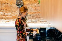 Vista laterale di felice giovane femmina in camicetta floreale mettere il pane in tostapane mentre si cucina la colazione in accogliente cucina al mattino — Foto stock