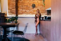 Sensuale giovane donna bionda in biancheria intima e camicetta floreale in piedi in cucina al mattino e smartphone di navigazione — Foto stock