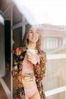 Glückliche attraktive junge Frau in stilvoller Dessous und farbenfroher Robe schaut zum Fenster, während sie den Morgen mit einer Tasse Kaffee und einem Smartphone zu Hause verbringt — Stockfoto