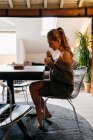 Vue latérale de la jeune femme déprimée en tenue décontractée assise à table avec une tasse de café dans les mains tout en prenant le petit déjeuner dans la solitude à la maison — Photo de stock
