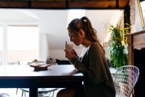Vue latérale de la jeune femme déprimée en tenue décontractée assise à table avec une tasse de café dans les mains tout en prenant le petit déjeuner dans la solitude à la maison — Photo de stock