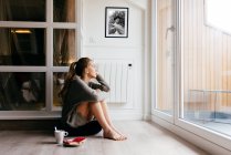 Side view of calm young female in casual wear looking at window thoughtfully while sitting on floor with cup of coffee and toasts during breakfast at home — Stock Photo