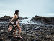 Side view of sensual pregnant woman looking away wearing long waving lingerie robe walking with bare belly on rocky coast in gloomy day — Stock Photo
