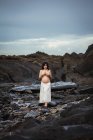 Dreamy topless brunette beautiful pregnant woman in maxi skirt standing on marvelous rocks with stream and covering breast looking at camera — Stock Photo