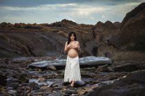 Dreamy topless brunette beautiful pregnant woman in maxi skirt standing on marvelous rocks with stream and covering breast looking at camera — Stock Photo