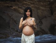 Verträumte brünette oben ohne schöne schwangere Frau in Maxi-Rock, die auf wunderbaren Felsen mit Strom steht und die Brust bedeckt und in die Kamera schaut — Stockfoto