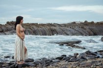 Dreamy topless brunette beautiful pregnant woman in maxi skirt standing on marvelous rocks with stream and covering breast looking away — Stock Photo