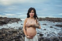 Мрійлива топлес брюнетка красива вагітна жінка в спідниці максі, що стоїть на чудових каменях з струмком і покриває груди, дивлячись на камеру — стокове фото