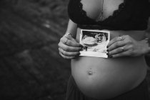 Татуйована стильна вагітна жінка в бюстгальтері тримає фотографію сонограми на животі, стоячи на величному морському узбережжі — стокове фото