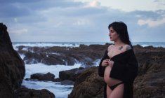Seitenansicht der glücklichen sinnlichen schwangeren Frau, die in einem langen wehenden Dessous-Gewand mit nacktem Bauch an der felsigen Küste an einem düsteren Tag wegschaut — Stockfoto