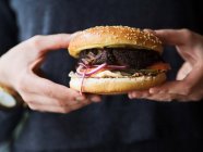 Ernte anonyme Person in schwarzer Kleidung hält klassische Burger mit Schnitzel und Gemüse mit Käse, während sie die Fast-Food-Industrie repräsentiert — Stockfoto