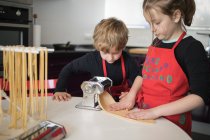 Uma menina com seu irmão usando máquina de massas enquanto prepara macarrão caseiro na cozinha doméstica — Fotografia de Stock