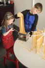 Зверху маленька дівчинка з старшим братом використовує макаронні машини, готуючи домашню локшину на домашній кухні — стокове фото