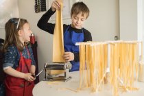 Маленькая девочка со старшим братом, использующая макароны во время приготовления домашней лапши на домашней кухне — стоковое фото
