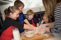Bambini allegri in grembiuli chef e donna delle colture in piedi intorno alla tavola e impasto impastare durante la preparazione di pasta artigianale — Foto stock