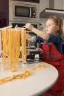 Grave menina pré-adolescente em avental vermelho pendurado macarrão cru no rack enquanto está à mesa com máquina de massas na cozinha — Fotografia de Stock