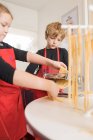 Una niña con hermano mayor que usa la máquina de pasta mientras prepara fideos caseros en la cocina casera - foto de stock