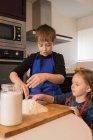 Menina bonito ajudando menino mais velho na preparação de massa, enquanto de pé juntos na mesa da cozinha com farinha — Fotografia de Stock
