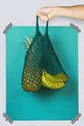 Unrecognizable person carrying net bag with ripe pineapple and bananas against blue turquoise rectangle during zero waste shopping — Stock Photo