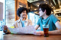 Von unten sehen sich junge homosexuelle Männer mit Navigationskarte und frischen Getränken lächelnd an, während sie beim romantischen Date am Cafétisch sitzen — Stockfoto