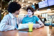 Hombres homosexuales jóvenes multiétnicos con mapa de navegación de dirección y bebidas frescas sonriendo mirándose mientras están sentados en la mesa de la cafetería durante una cita romántica - foto de stock