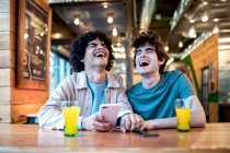 Багатонаціональні молоді гомосексуальні чоловіки переглядають соціальні медіа на смартфоні і мають свіжі напої, посміхаючись закритими очима, сидячи за столом кафе під час романтичного побачення — стокове фото